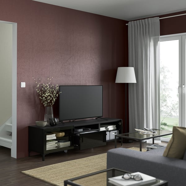 Modern living room: maroon walls, BESTÅ tv bench, books, flowers, remote on glass table in front of grey sofa.
