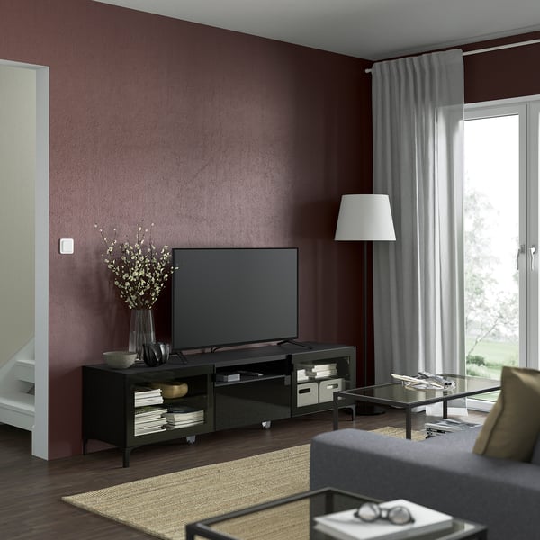 Modern living room with maroon walls, black BESTÅ tv stand, flowers, lamp, glass table, natural light.