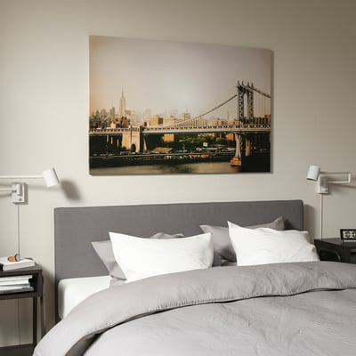 Large canvas print of manhattan bridge on hotel wall, above grey upholstered bed with white pillows. BJÖRKSTA frame highlights consistent decor.