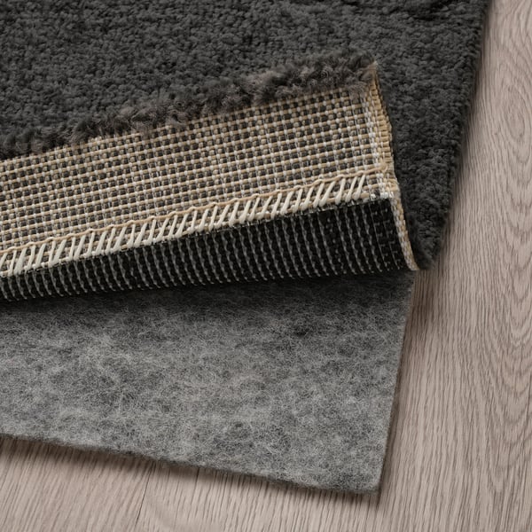 A dark grey rug with curly yarn pattern lies on a wooden floor, showing an anti-slip pad underneath.