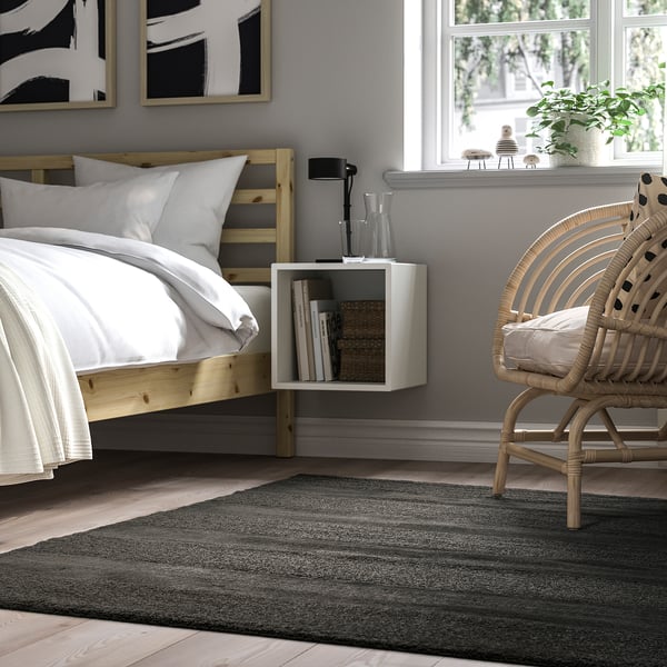 Bedroom with LANDSVÄG rug, wooden bed, wicker chair, side table, lamp, plant, window, artwork.