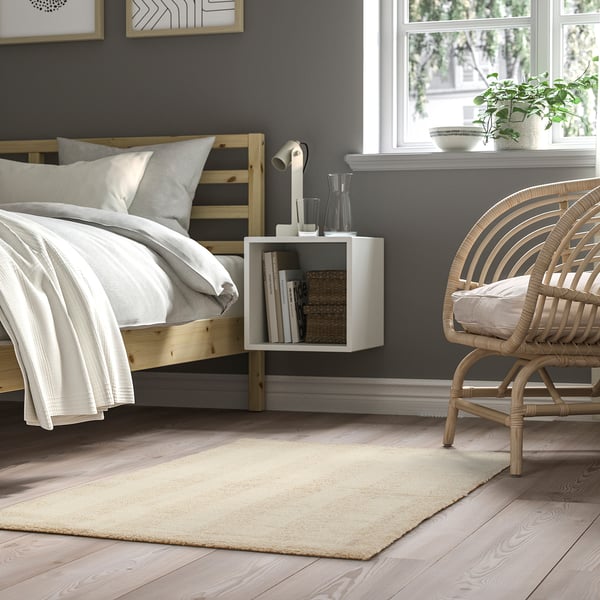 Bedroom with grey LANDSVÄG rug, wooden bed, white side table, wicker chair, and wall art near window.