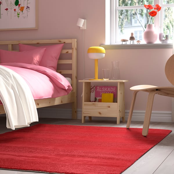 Bright pink bedroom with wooden furniture, featuring a red IKEA LANDSVÄG rug, a yellow lamp, and a wooden bed.