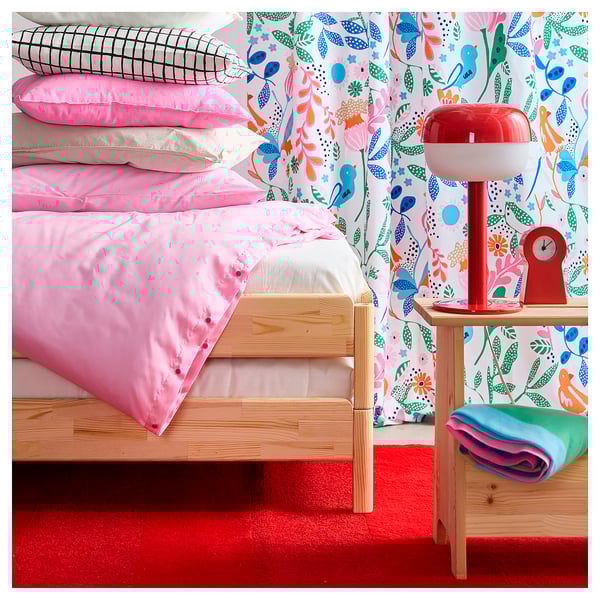 Bright, red area rug under a wooden bed with colourful pillows and a floral wallpaper.