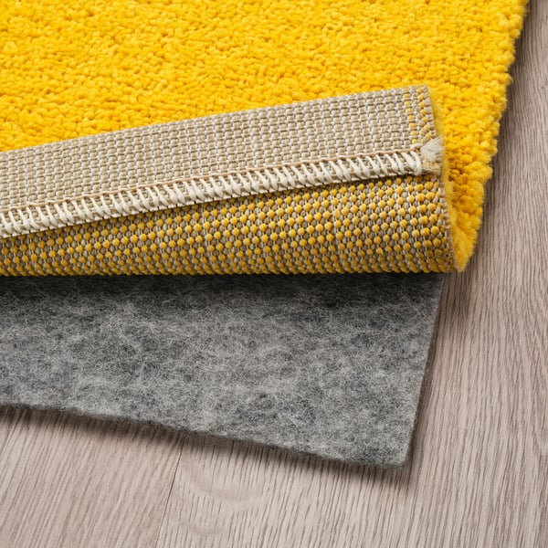 Bright yellow soft rug, folded to show layers. A grey non-slip rug underlay is visible underneath. Ideal for living rooms, adding warmth and comfort.
