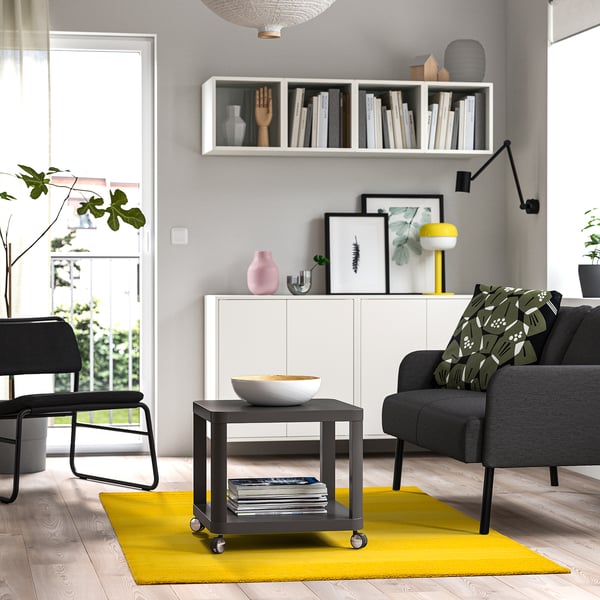 Modern living room with LANDSVÄG yellow rug, grey sofa, black coffee table, and white shelves filled with books and decor.
