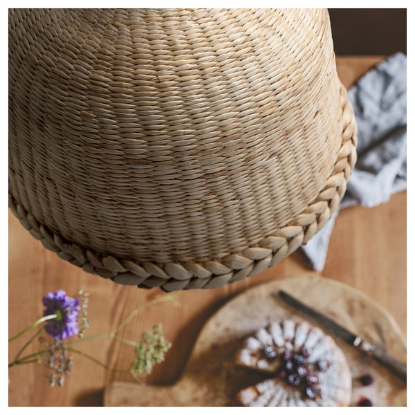 Close-up of an upside-down beige hand-braided banana fibre lampshade, round with intricate patterns, on a wooden surface with a colourful flower.