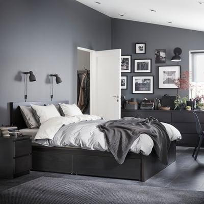 Modern grey bedroom with high-bed, bedside lamps, white pillows, dark dresser with plants, black frames on wall, coats on door.
