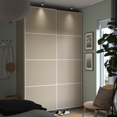 A modern bedroom a tall beige PAX wardrobe with sliding doors, soft lighting, a side table with a cushion, and a potted plant.