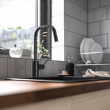 Moderne Küchenspüle mit schwarzer ÄLMAREN-Armatur, wasser- und energiesparend, hoher Auslauf für einfache Reinigung, Fensternähe mit Pflanzen und Geschirr.