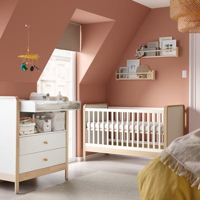 Babyzimmer mit weißem Kinderbett und Kommode aus Holz, rosa Wänden, gelbem Hängemobile, Regalen mit Spielzeug, gemütlichem Teppich.