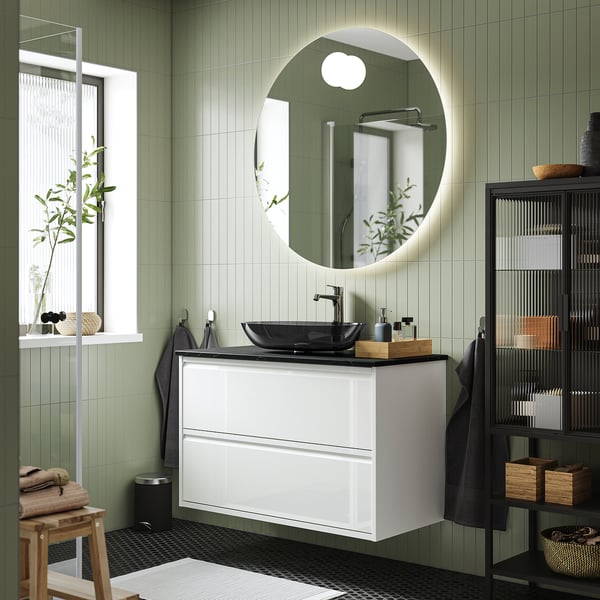Modernes ÄNGSJÖN-Badezimmer: weißer Schrank, runder Spiegel, dunkles Waschbecken; Wandmontage mit grün gefliesten Wänden und schwarzen Armaturen.