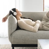 Frau entspannt sich auf grauem ÄPPLARYD-Sofa, trägt Kopfhörer, beige Pullover und Leggings.