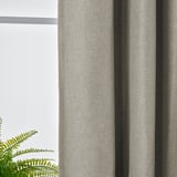 Nahaufnahme von strukturierten, raumverdunkelnden Vorhängen in Hellgrau, die teilweise zurückgezogen sind und grüne Farnblätter in der Nähe eines Fensters zum Vorschein bringen.
