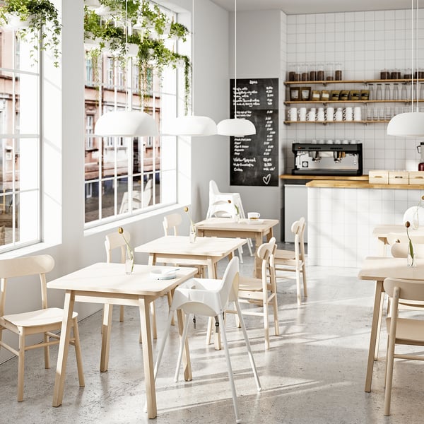 Modernes Café mit weißen Tischen, Stühlen und Pendelleuchten; Große Fenster, üppige Pflanzen, Kreide-Menütafel und Kaffeezubehör.