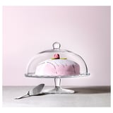 Tortenständer aus Glas mit durchsichtigem Kuppeldeckel, der rosa Kuchen hält. Daneben liegt ein silberner Servierspatel.