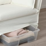 Durchsichtige Aufbewahrungsbox aus Kunststoff unter einem weißen Sofa, in der gefaltete Decken und Kissen aufbewahrt werden.