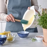 Person, die mit einem Spatel Flüssigkeit aus einer Schüssel gießt. Schüsseln mit Zutaten stehen auf dem Tisch.