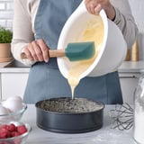 Eine Person gießt Teig aus einer weißen Schüssel mit einem grünen Spatel in eine runde Pfanne, umgeben von Backzutaten auf einer Küchentheke.