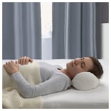 Eine Person, die ein BARRBERBERIS Nackenkissen auf einem Bett benutzt. Das Kissen ist weiß, besteht aus Baumwolle und Entenfedern und hat ein klassisches Design für die Nackenstütze.