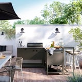 Moderne Außeneinrichtung mit BÅTSKÄR-Küche aus schwarzem Stahl mit Arbeitsplatte, Grill, Hockern und Möbeln vor einer weißen Wand mit Lichtern.