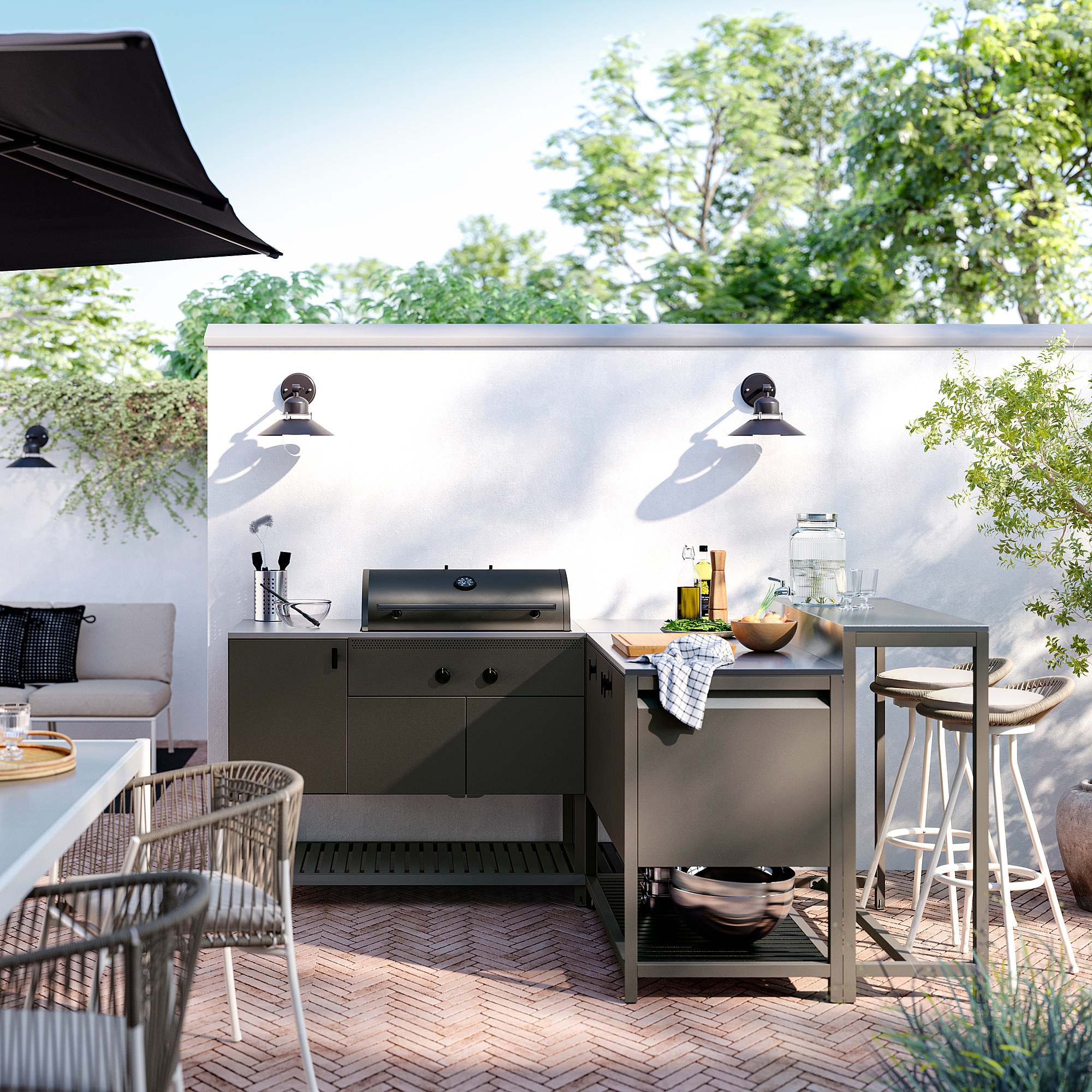 Moderne Außeneinrichtung mit BÅTSKÄR-Küche aus schwarzem Stahl mit Arbeitsplatte, Grill, Hockern und Möbeln vor einer weißen Wand mit Lichtern.