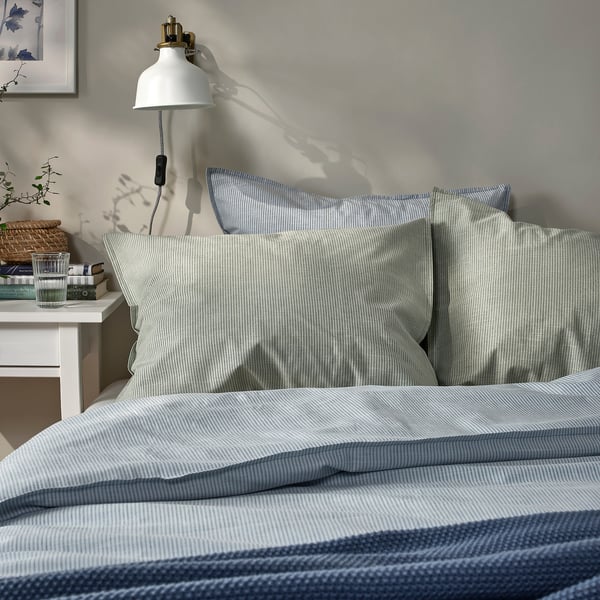 Ordentlich gemachtes Bett mit hellblau gestreiften Kissenbezügen, Nachttisch, Lampe, Büchern, Glas; Gemütliches, kompaktes Schlafzimmer.