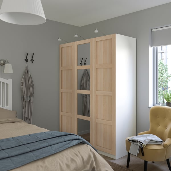 Ein Schlafzimmer mit hellem BERGSBO-Kleiderschrank, dessen Spiegeltür den Raum widerspiegelt, neben einem großen Bett und einem gelben Sessel.