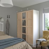 Modernes Schlafzimmer mit großem BERGSBO-Holzschrank und Spiegeltür, positioniert zwischen Bett und Fenster. Graue Wände, gelber Stuhl und hellbraune Bettwäsche.