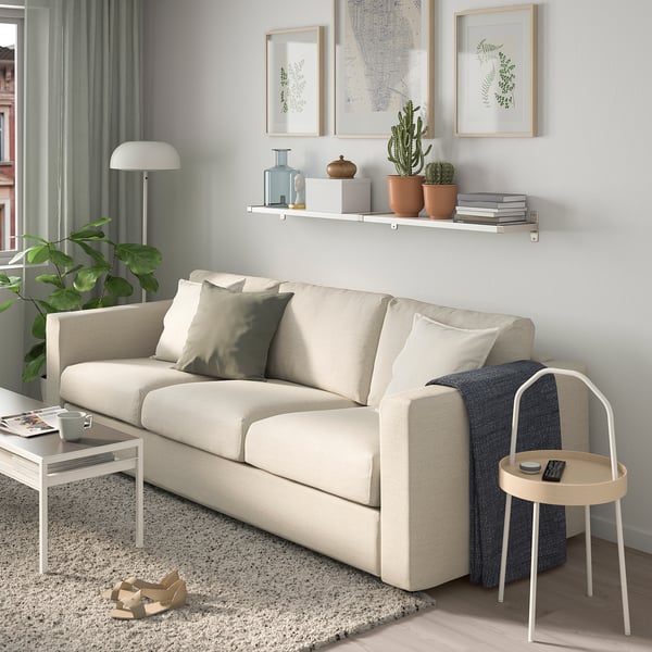 Modernes Wohnzimmer mit beigem Sofa, Holzboden, grauen Vorhängen, Wandkunst und Pflanzen. Weißes Regal mit Büchern und Pflanzen.