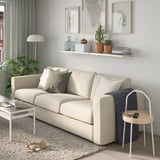 Modernes Wohnzimmer mit beigem Sofa, Holzboden, grauen Vorhängen, Wandkunst und Pflanzen. Weißes Regal mit Büchern und Pflanzen.
