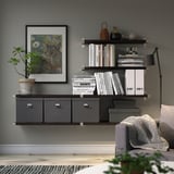 Modernes Wohnzimmer mit grauen BERGSHULT-Regalen mit Büchern, Vasen und Katzenkunst über einem grauen Sofa mit gemütlichem Überwurf.