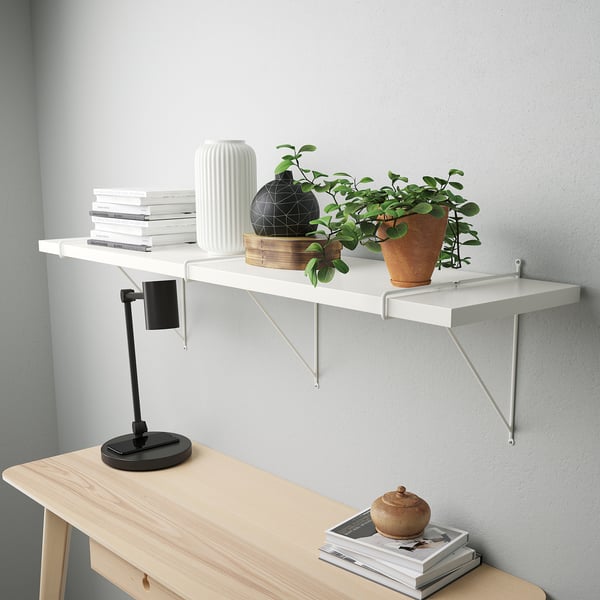 Ein Wandregal mit Büchern und Pflanzen. Es hat eine abgeschrägte Kante und weiße Klammern. Darunter befindet sich ein Schreibtisch mit einer Lampe und weiteren Büchern.