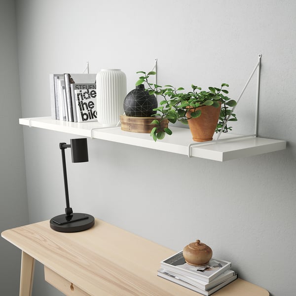 Modernes weißes Wandregal mit abgeschrägter Kante, für Bücher, Topfpflanze, schwarze Vase. Darunter ein Konsolentisch aus hellem Holz mit Lampe und Zeitschriften.