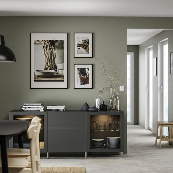 Modernes Wohnzimmer mit salbeigrünen Wänden. Schwarzes BESTÅ Sideboard mit Glastüren, zwei Schubladen und Dekorationsartikeln.