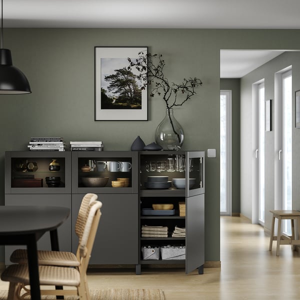 Grauer BESTÅ-Schrank mit Glastüren, Küchenutensilien, schwarzer Vase, Holzstuhl, grüner Flur.