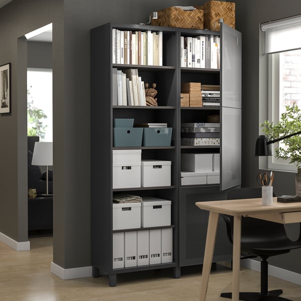 Schwarzer BESTÅ-Aufbewahrungsschrank mit offenen und geschlossenen Regalen für Bücher, Schachteln und Dekorationsartikel in einem Home-Office-Setup.