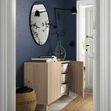 Flur mit BESTÅ Sideboard aus hellem Holz, offene Türen, die Staukästen freigeben. Oben, runder Spiegel und Wandleuchte an blauer Wand.