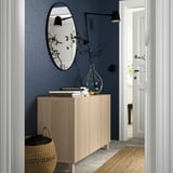BESTÅ Sideboard aus hellem Holz mit zwei Türen, dunkelblaue Wand, schwarzer ovaler Spiegel, gestapelte Bücher, Vase, Korb, heller Raumblick, schwarze Wandlampen.
