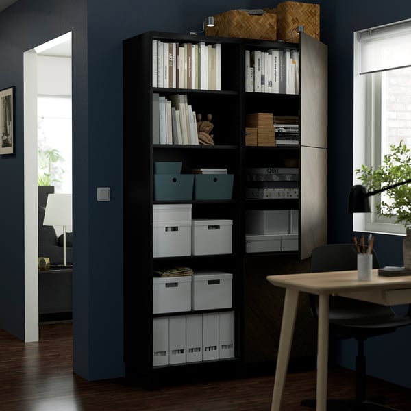 Dunkler Holzschrank mit mehreren Regalen, in dem verschiedene Bücher und Behälter aufbewahrt werden, neben einem Holztisch mit Stuhl.