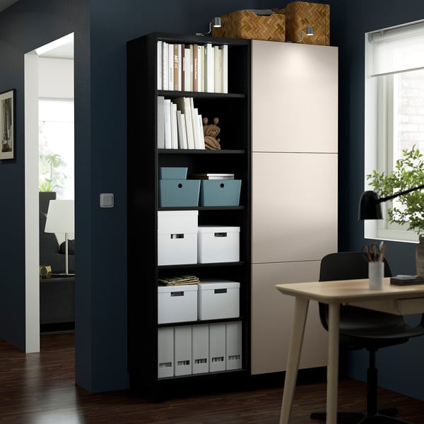 Ein hoher schwarzer BESTÅ-Schrank mit weißen Türen in einem modernen Raum, in dem Aufbewahrungsboxen und Bücher aufbewahrt werden, neben einem Schreibtisch und einem Stuhl.