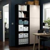 Ein hoher schwarzer BESTÅ-Schrank mit weißen Türen in einem modernen Raum, in dem Aufbewahrungsboxen und Bücher aufbewahrt werden, neben einem Schreibtisch und einem Stuhl.