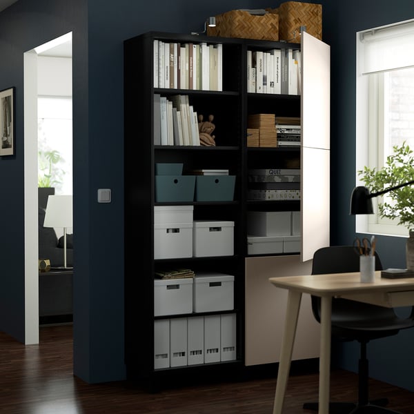 Schwarze Regale mit Büchern, Kisten und Dekoration vor einer dunkelblauen Wand, in der Nähe eines Arbeitsbereichs mit Schreibtisch und Stuhl.