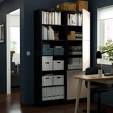 Schwarze Regale mit Büchern, Kisten und Dekoration vor einer dunkelblauen Wand, in der Nähe eines Arbeitsbereichs mit Schreibtisch und Stuhl.