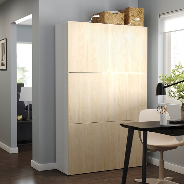 Stellen Sie sich einen hellen, holzfarbenen BESTÅ-Aufbewahrungsschrank in einem modernen Wohnraum vor, einschließlich eines Schreibtisches mit Lampe und Stuhl.