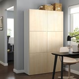 Stellen Sie sich einen hellen, holzfarbenen BESTÅ-Aufbewahrungsschrank in einem modernen Wohnraum vor, einschließlich eines Schreibtisches mit Lampe und Stuhl.
