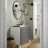 Moderner Flur: BESTÅ Sideboard in Grau, großer runder Spiegel, Wandlampen, Vase, Bücher, geflochtener Korb, der in einen hellen Raum führt.