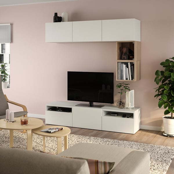 Modernes Wohnzimmer mit weißem BESTÅ-TV, Flachbildfernseher, Pflanzen, grauen Vorhängen, Couchtischen aus hellem Holz und beigem Sofa auf einem gemusterten Teppich.