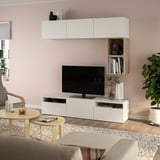 Modernes Wohnzimmer mit weißem BESTÅ-TV, Flachbildfernseher, Pflanzen, grauen Vorhängen, Couchtischen aus hellem Holz und beigem Sofa auf einem gemusterten Teppich.