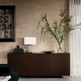 Modernes Wohnzimmer mit dunkelbraunem BESTÅ-Schrank, grünem Stuhl, weißer Lampe, hoher Pflanze in Glasvase und gerahmtem Kunstwerk an strukturierter Wand.